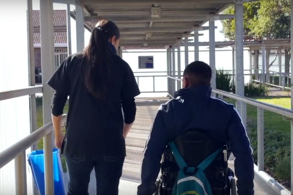 A man using a wheelchair travels away from the camera, carrying a backpack on the back of his chair. A woman walks next to him.