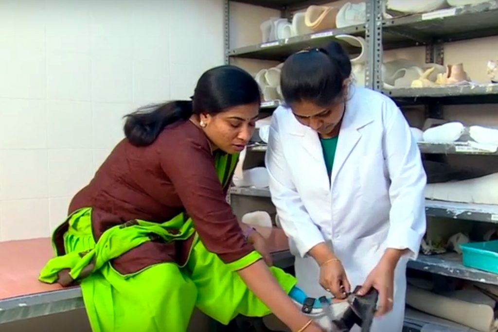 A woman sits on a high bench, raising her leg so a female service provider in a white coat can examine her foot orthosis.
